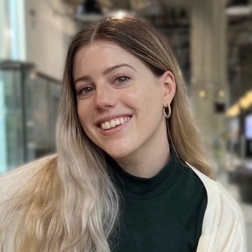 A smiling woman with long, wavy blonde hair and hoop earrings sits in an indoor setting. She is wearing a dark green top and a light-colored cardigan. The background, slightly blurred, shows industrial-style decor with hanging lights. She is part of The Representation Project's Board of Directors.
